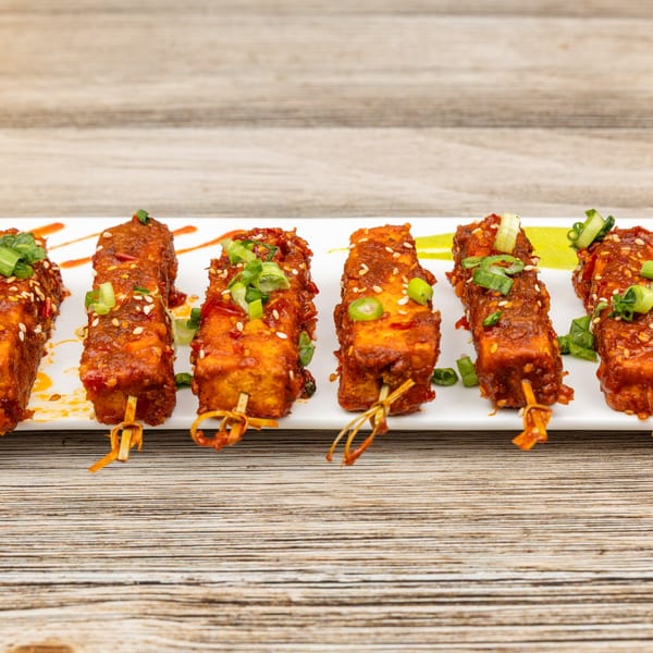 Six Indian paneer tikka skewers, coated in red sauce, sesame seeds, and green onions on a white plate.