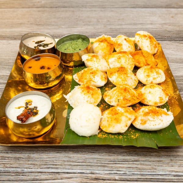 Indian idli with three chutneys in golden bowls on a banana leaf and gold tray.