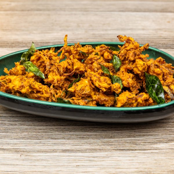 A close-up of golden-brown Indian pakora, garnished with fresh green curry leaves, in a dark green bowl.