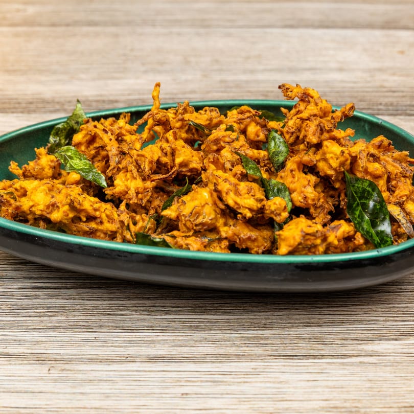 A close-up of golden-brown Indian pakora, garnished with fresh green curry leaves, in a dark green bowl.