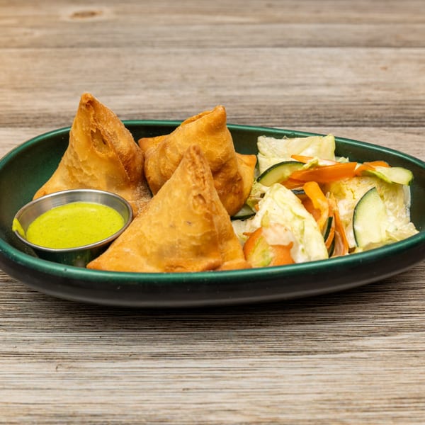 Four golden-brown samosas with green dipping sauce and a side salad in a dark green bowl.