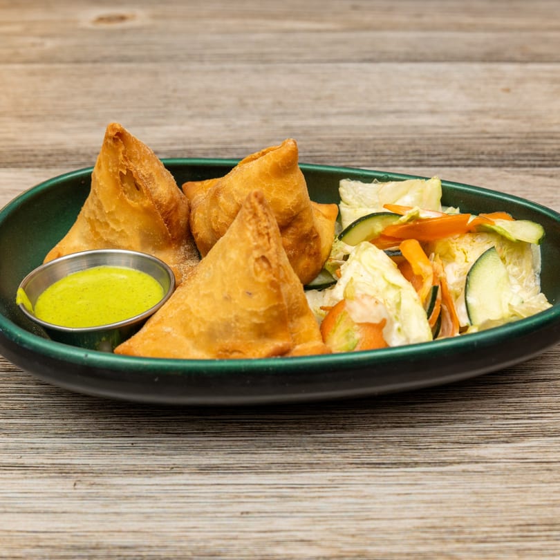 Four golden-brown samosas with green dipping sauce and a side salad in a dark green bowl.
